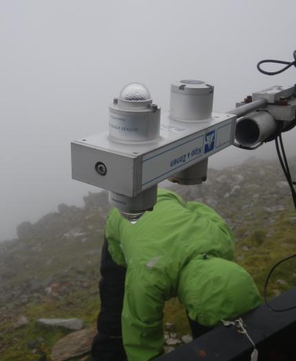A person in a green jacket adjusts scientific monitoring equipment in a foggy, mountainous setting.